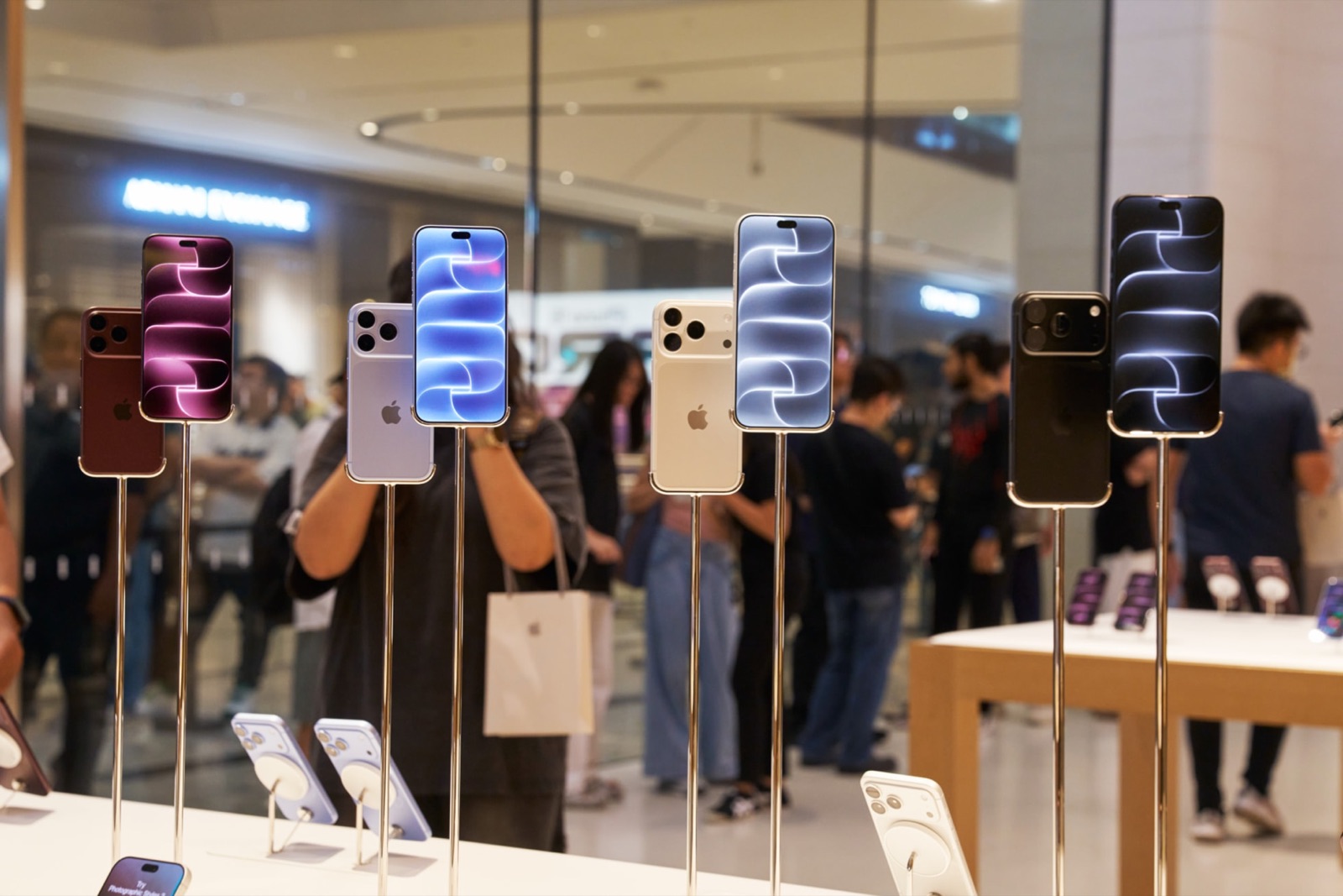 Display iPhone 18 Pro units in burgundy, glacier, silver and black stand on chrome mounts on a table inside Apple The Exchange TRX, with shoppers and a paper Apple bag blurred behind the glass.
