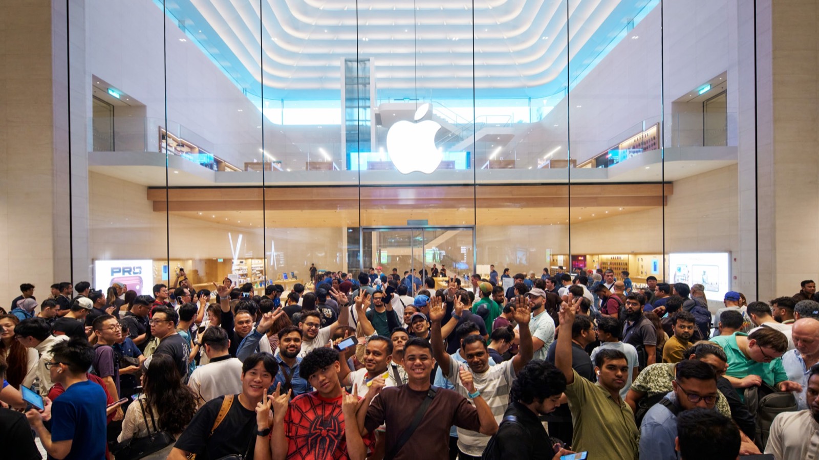 A crowd of customers fills the atrium in front of the glass facade of Apple The Exchange TRX in Kuala Lumpur on iPhone 18 Pro launch day, many raising their hands and holding phones, with the illuminated Apple logo and the curved white ceiling of the store behind them.